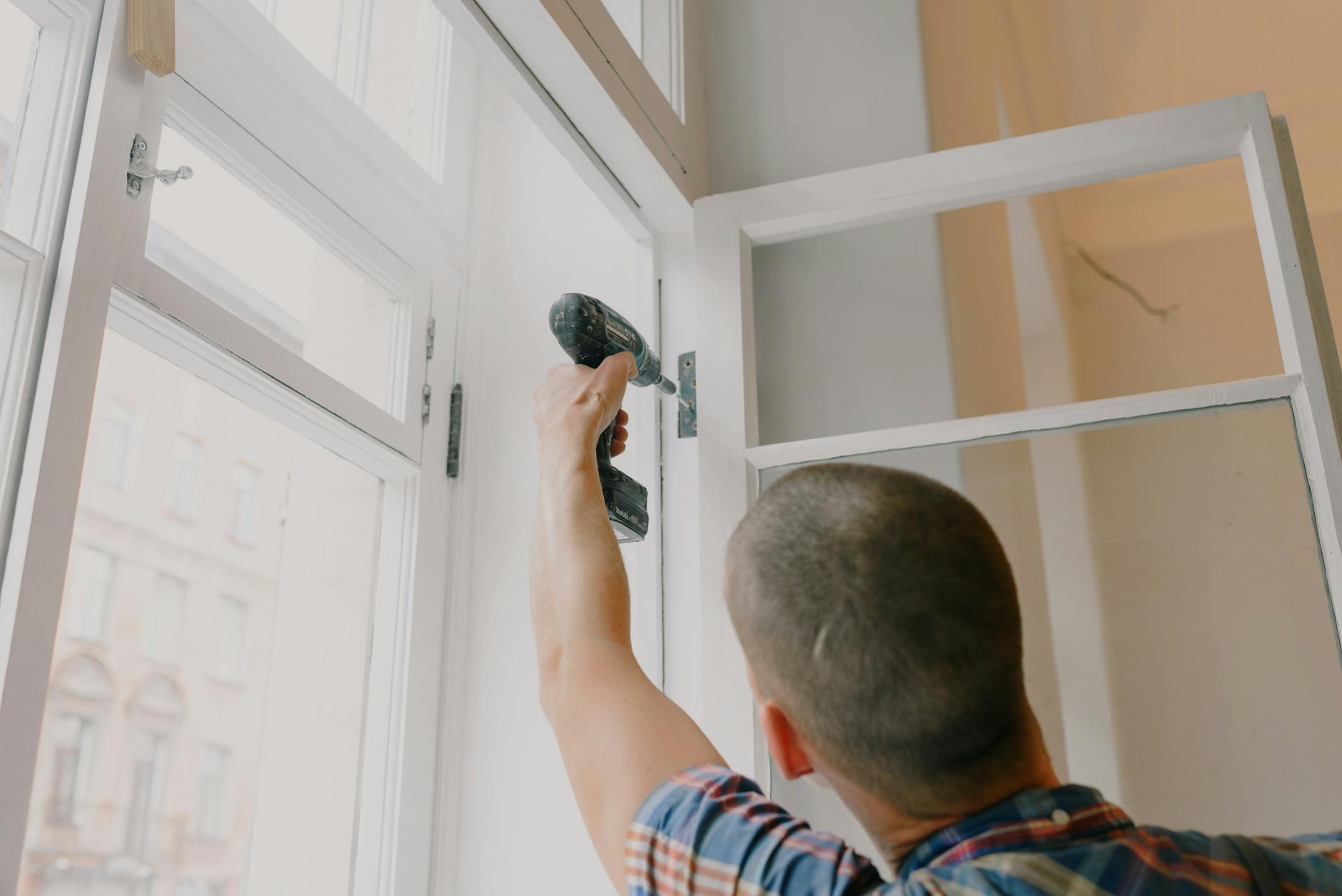 A handyman works with a drill installing window hinges indoors.